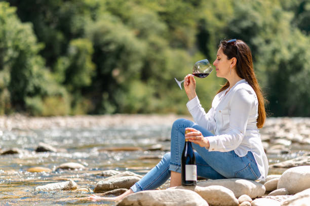istock Attractive woman drinking red wine sitting on a rock in the riverside. 1321494094