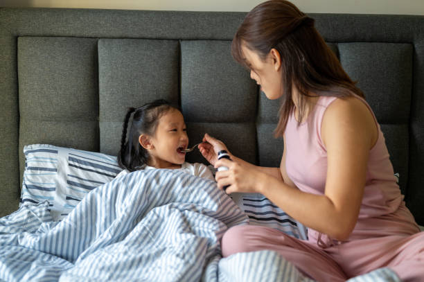 una mamma asiatica che versa la medicina dello sciroppo per la tosse in un cucchiaio chiaro a sua figlia - cold remedy foto e immagini stock