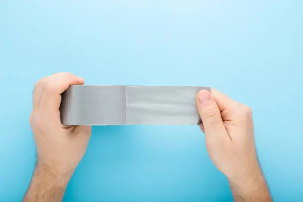 Young adult man hands stretching gray adhesive tape on light blue table background. Pastel color. Closeup. Point of view shot. Top down view.