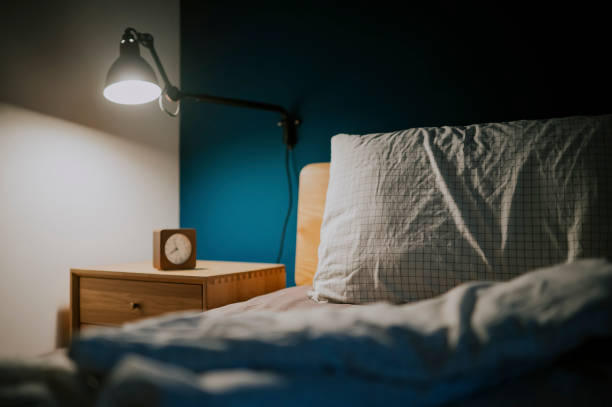 青い壁が付いているベッドの横の夜のテーブルの上の時計と電気ランプによって照らされた夜の寝室 - pillow headboard wall bedroom ストックフォトと画像