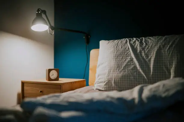 Photo of bedroom at night illuminated by electric lamp with clock on night table beside the bed with blue wall