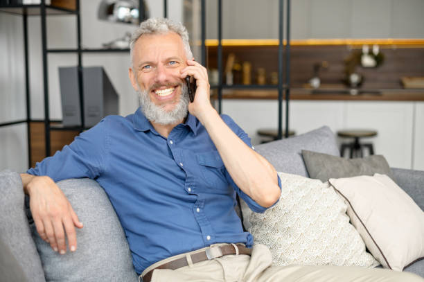 istock Senior bearded guy sitting down on the comfortable couch and talking on the smartphone 1321505051