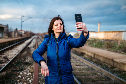Young beautiful smiling Caucasian woman making selfies on railway track while waiting for a train.