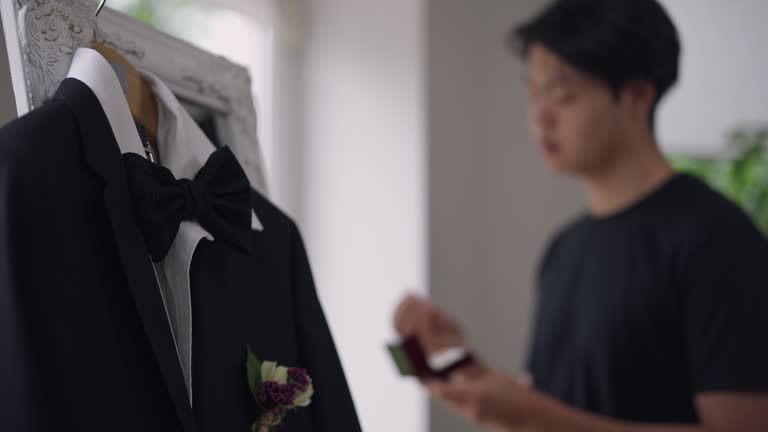 Close-up of wedding costume with blurred Asian man looking at wedding rings at background. Blazer with boutonniere and bow tie hanging on mirror as groom admiring jewelry in the morning. Marriage.