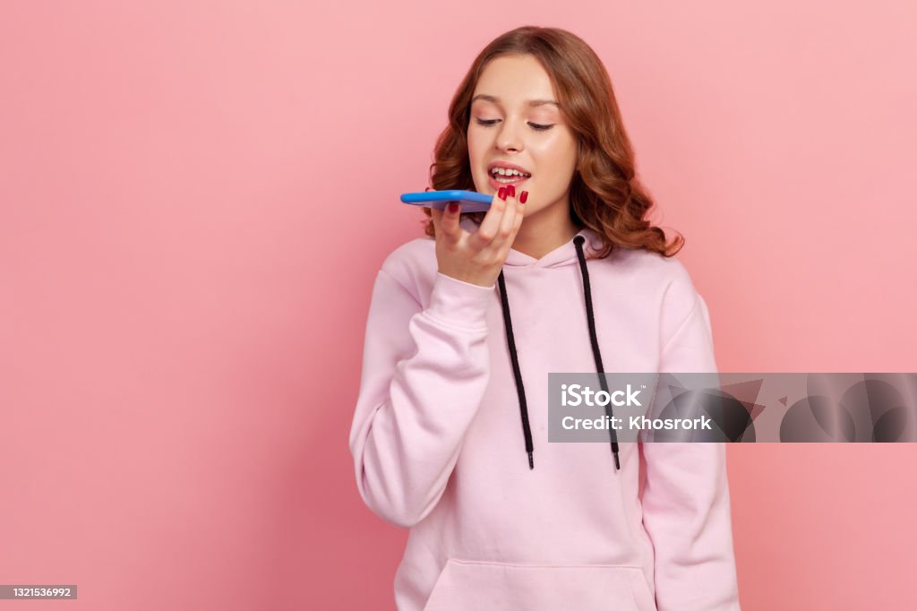 Portrait of brunette teen girl in hoodie talking to smartphone, using virtual assistant, ai intelligent technology, voice application, audio message Portrait of brunette teen girl in hoodie talking to smartphone, using virtual assistant, ai intelligent technology, voice application, audio message. Indoor studio shot isolated on pink background Talking Stock Photo