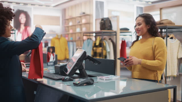 Clothing Store Checkout Cashier Counter: Woman and Male Retail Sales Managers Accept NFC Smartphone Payment from a Young Stylish Female Customer for Clothes and Pack Them in a Recyclable Bag.