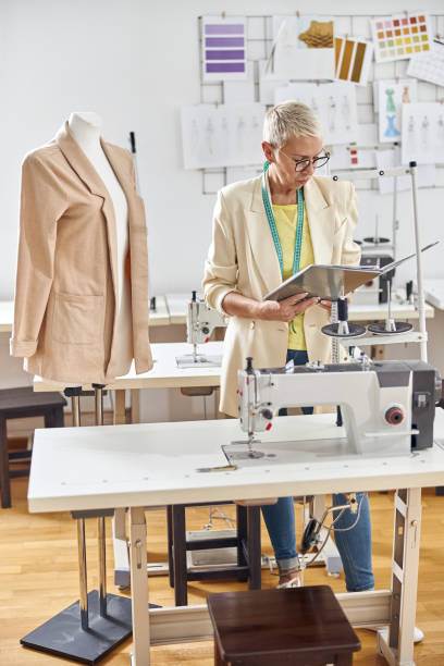 istock Middle aged fashion designer looks at open folder standing by table with sewing machine 1321540893