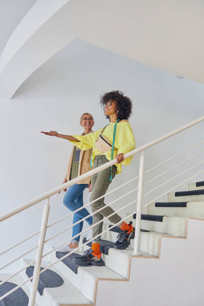 African-American dressmaker points on something in front to mature colleague going down on steps