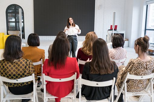 Rear view of unidentified people listening to coach having speech at business training. A group of women attending  seminar in conference hall. Life long learning