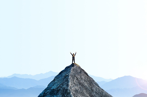 A businesswoman standing on the peak of a mountain top raises her arms as she celebrates attaining her goal.