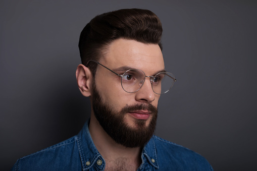 Confident smart modern bearded man in casual denim clothes and eyewear is posing over gray background in the studio