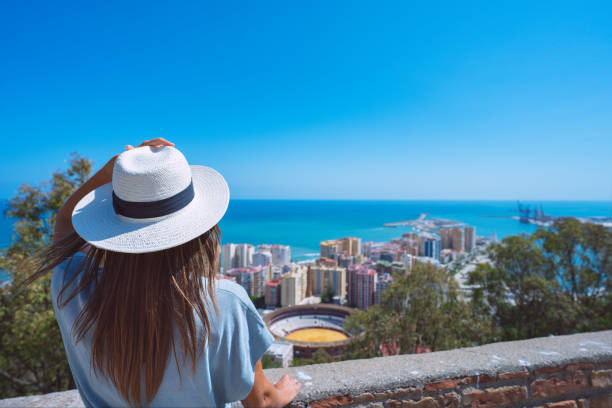 vista posteriore della giovane donna che viaggia con cappello bianco in piedi nel punto panoramico del castello di gibralfaro e guardando il paesaggio urbano di malaga. vacanze estive in spagna - provincia di málaga foto e immagini stock