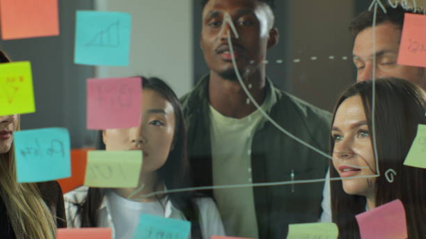 istock Diverse coworkers group analyzing corporate strategy drawing chart on glass wall with sticky notes. Multiracial business team people designers or marketing experts brainstorm at business meeting. 1321664679