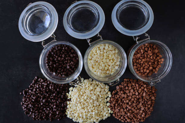 Image of row of transparent, open lid, full jars besides row of individual piles of milk, white and dark chocolate chips on black background, elevated view Stock photo showing an elevated view studio shot on a dark black surface of lidded jars filled with white, milk and dark chocolate chips. chocolate white chocolate chocolate chip white stock pictures, royalty-free photos & images