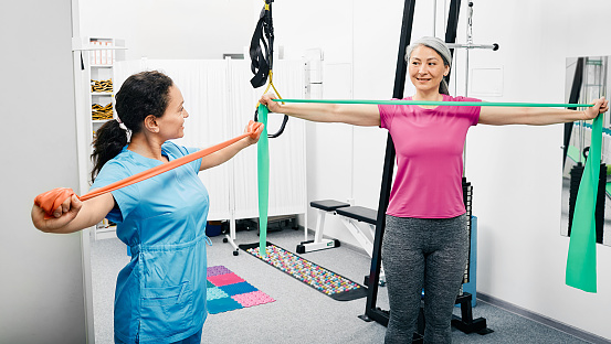 Rehabilitation therapist shows correct execution of exercise with a resistance band. Adult woman uses an elastic resistance band for training her arms