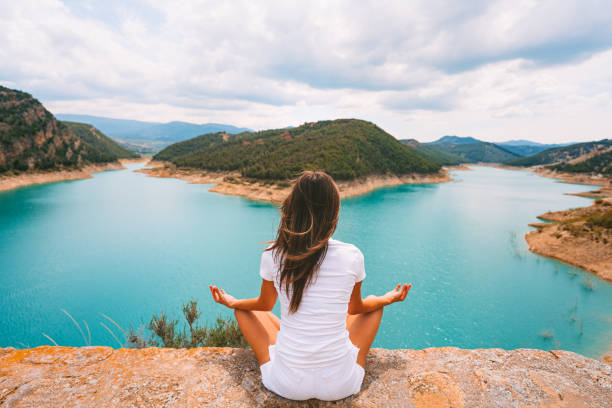 Healthy young woman in white clothes meditating or practicing yoga in the nature. Woman sitting in yoga position, easy pose or sukhasana with mudra with beautiful blue lake view. Relaxation, harmony. Healthy young woman in white clothes meditating or practicing yoga in the nature. Woman sitting in yoga position, easy pose or sukhasana with mudra with beautiful blue lake view. High quality photo sukhasana stock pictures, royalty-free photos & images