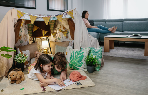 Children playing camping at home while their mother watches TV sitting on the sofa