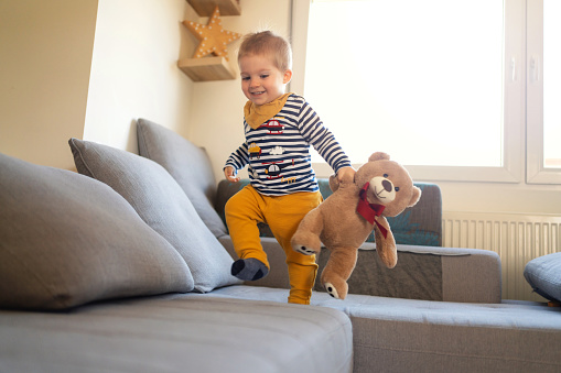 Happy Caucasian baby boy having fun while walking on the sofa with his teddy bear.