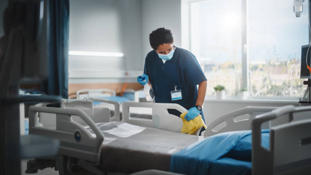ziekenhuisafdeling: professionele zwarte verpleegster die gezichtsmasker draagt, het bed afveegt, de kamer schoonmaakt nadat covid-19-patiënten herstellen. desinfectie, sterilisatie, ontsmettingskliniek na coronavirus geïnfecteerde mensen - ziekenhuis stockfoto's en -beelden