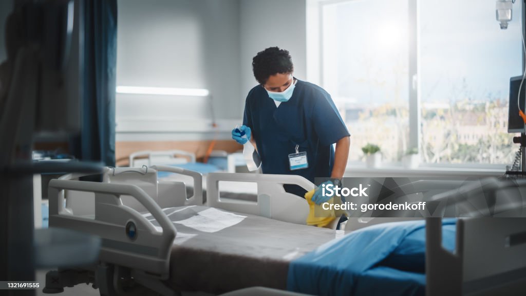 Hospital Ward: Professional Black Nurse Wearing Face Mask, Wiping the Bed, Cleaning Room After Covid-19 Patients Recover. Disinfection, Sterilizing, Sanitizing Clinic after Coronavirus Infected People Hospital Stock Photo
