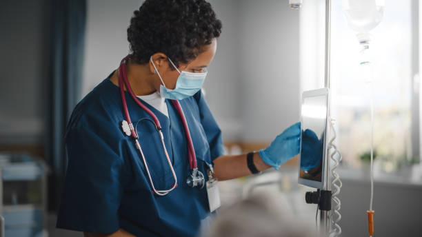 istock Hospital Ward: Professional Black Head Nurse Wearing Face Mask Does Checkup of Patient's Vitals, Checking Heart Rate Computer, Intravenous or Iv Fluids Drip Bag. Caring Nurse Monitors Person Recovery 1321691575