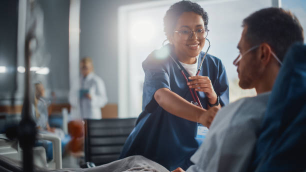 salle d’hôpital: amical black head nurse utilise stéthoscope pour écouter les battements de cœur et les poumons du patient masculin en convalescence se reposant dans son lit, fait un bilan. l’homme se bien après une chirurgie réussie - human cardiopulmonary system photos et images de collection