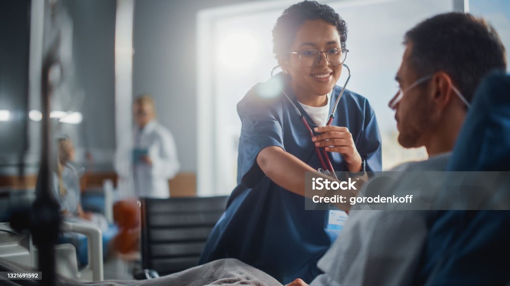 Hospital Ward: Friendly Black Head Nurse Uses Stethoscope to Listen to Heartbeat and Lungs of Recovering Male Patient Resting in Bed, Does Checkup. Man Getting well after Successful Surgery Patient Stock Photo