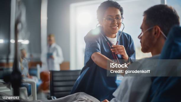 Hospital Ward Friendly Black Head Nurse Uses Stethoscope To Listen To Heartbeat And Lungs Of Recovering Male Patient Resting In Bed Does Checkup Man Getting Well After Successful Surgery Stock Photo - Download Image Now