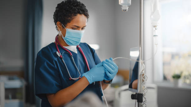 istock Hospital Ward: Professional Black Head Nurse Wearing Face Mask Does Checkup of Patient's Vitals, Checking Heart Rate Computer, Intravenous or Iv Fluids Drip Bag. Caring Nurse Monitors Person Recovery 1321691596