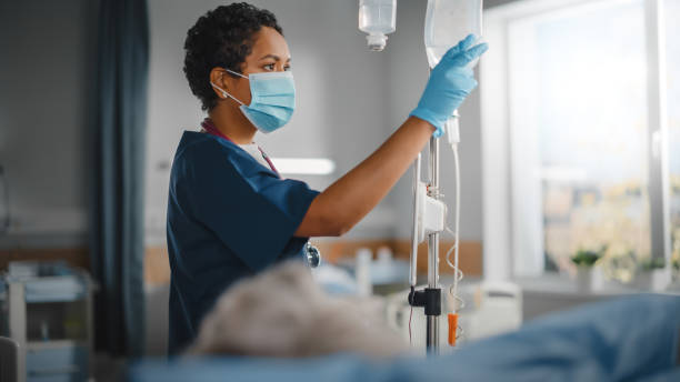 hospital ward: professional black head nurse wearing face mask does checkup of patient's vitals, checking heart rate computer, intravenous or iv fluids drip bag. caring nurse monitors person recovery - hospital imagens e fotografias de stock