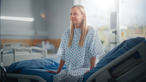 modern hospital ward: beautiful caucasian female patient resting on a bed, fully recovering after successful surgery, sickness or coronavirus she smiles. behind her window with a beautiful sunny view - cirurgia imagens e fotografias de stock