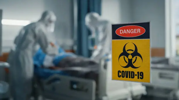 Photo of Hospital Coronavirus Emergency Department Ward: Doctors wearing Coveralls, Face Masks Treat, Cure and Save Lives of Patients. Focus on Biohazard Sign on Door, Background Blurred Out of Focus