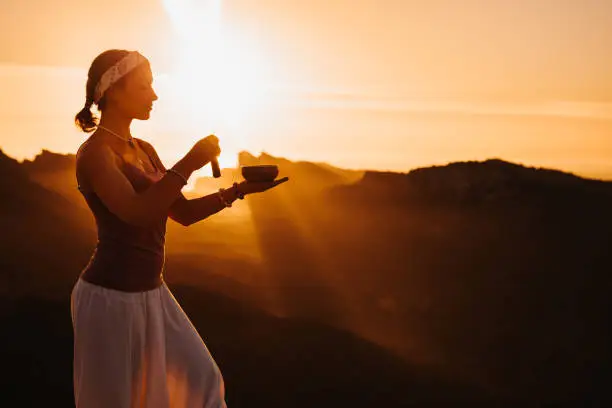 Beautiful woman doing meditation using a Tibetan singing bowl during the sunrise in a mountainous landscape in Majorca. Color editing with slightly grain. Part of a series.