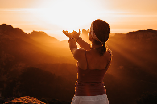 Silhouette of a  young woman in spiritual pose holding the light and sunbeams with both hands in front of mountains. Creative color editing. Selective focus on the hands. Part of a spiritual series.