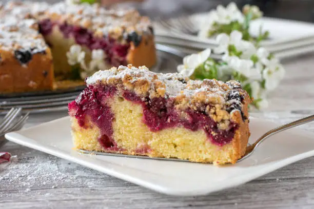 A Piece of Cherry crumble cake on a plate on white wooden table with closeup view and whole cake in the background with cherry blossoms decoration