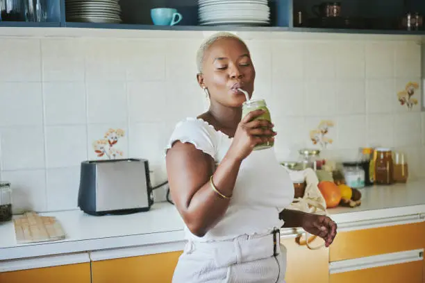 Young African woman standing with her eyes closed in her kitchen at home and drinking an organic smoothie with a reusable straw