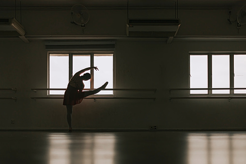 in silhouette asian chinese teenage girl practicing ballet dance raise her leg on barre in the studio