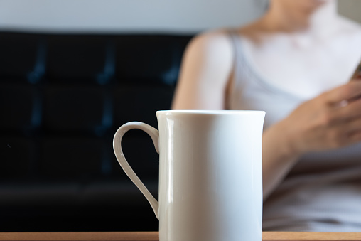 A woman looking at her smartphone while drinking coffee in her living room in the morning.\nA person operating a smartphone in the background with coffee as the main ingredient. Back blur. Selective focus.