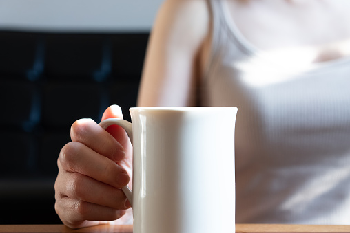 A woman relaxing with a cup of coffee in the living room of her house in the morning.\nThe hand of the person holding the coffee. Back bokeh. Selective focus.