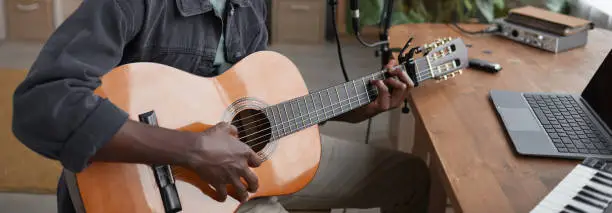 Portrait of young African-American man playing guitar and looking at camera while sitting by microphone in home recording studio, copy space