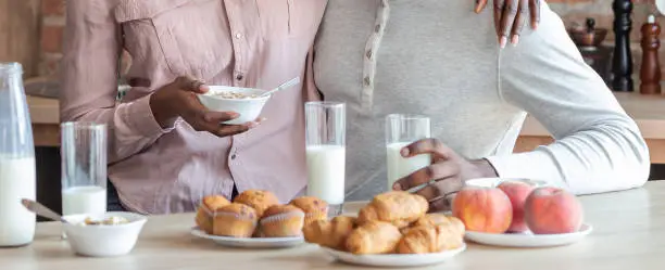 African young married couple having breakfast together at kitchen, woman sitting on man lap and hugging him, drinking milk and eating pastry, copy space