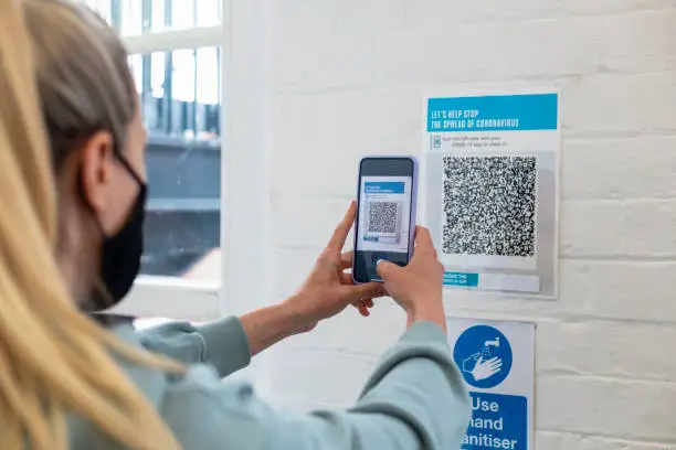 Over the shoulder view of a mid-adult woman wearing a protective face mask standing in a vaccine waiting room, scanning a check-in barcode, in preparation of getting her COVID-19 vaccine.