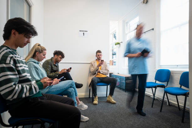 Waiting With Fellow Customers People sitting in a vaccine waiting room, they are waiting to get their COVID-19 vaccine. They are all using their mobile phones. There is a medical professional walking through the room, who is blurred in motion. waiting room stock pictures, royalty-free photos & images