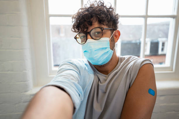 Today is the Day! Selfie shot of a man sitting in the doctor's office taking a selfie with his arm which has a blue plaster on it, after getting the COVID-19 vaccine. rolled up sleeves stock pictures, royalty-free photos & images