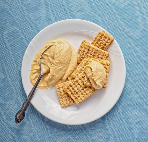 Smooth and creamy hummus on plate with woven wheat crackers. Healthy eating hummus is a good snack paired with whole grain wheat crackers. Snack food on small white plate with spoon on a blue tablecloth background.