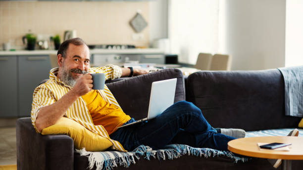 istock Senior man with gray beard looking at camera and toasting with cup of tea while working on laptop computer placed on his lap sitting on comfortable sofa at home 1321750096