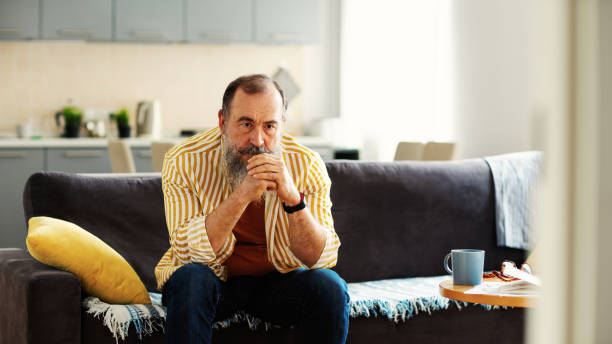 istock Depressed senior man wirh gray beard sitting on sofa at home being deep in thought. Frustrated male thinking sadly with his hands clasped 1321750106