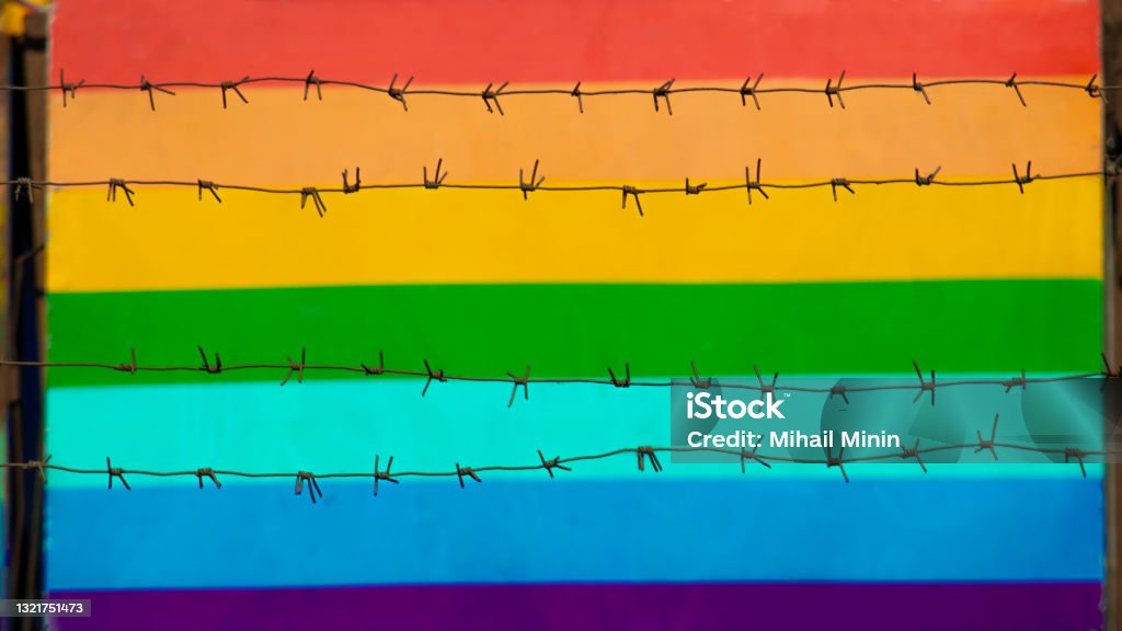 multicolored gay community flag behind barbed wire multicolored flag of gay community behind barbed wire in blurred bokeh Homophobia Stock Photo