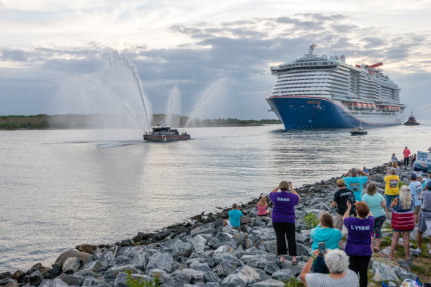 Cape Canaveral, Florida, USA - June 4, 2021: The cruise ship Carnival Mardi Gras arrives  in the USA at Port Canaveral. It is escorted by a fireboat shooting water cannons. A crowd watches from the shore at Jetty Park, some carrying welcoming signs.