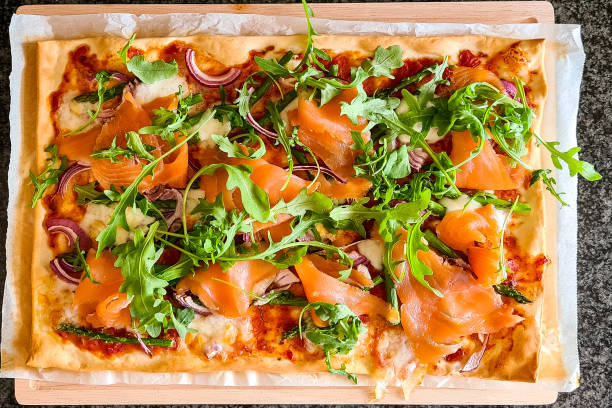 Studio photo of wooden plank with a tarte flambée or flammkuchen on baking paper. The flammkuchen is covered with goat cheese, green asparagus, onion, rocket  and smoked salmon.